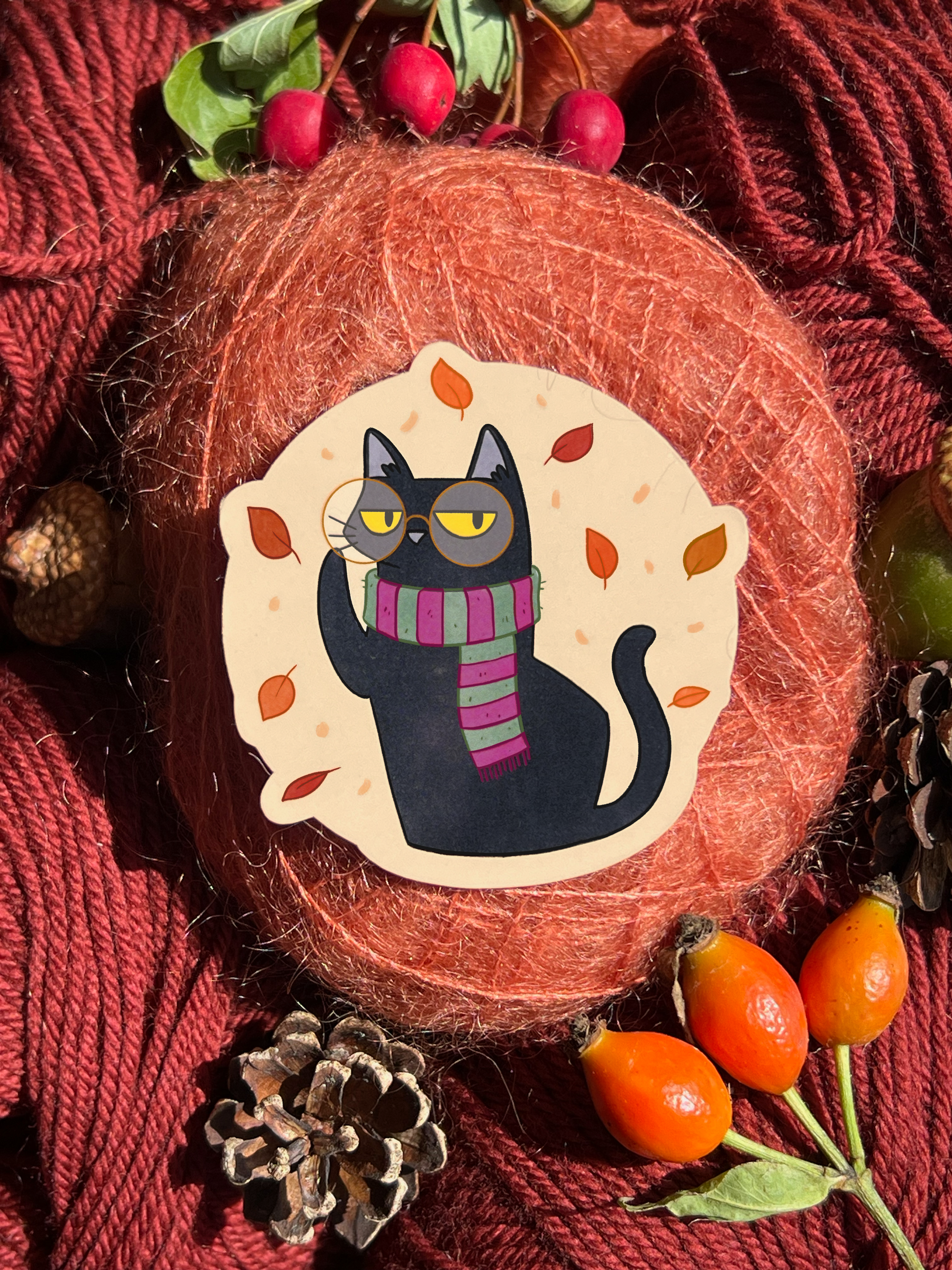 Autumn Blackgato Sticker product featurering illustration of black cat in autumn themed clothing. Cosy background of red yarn, pinecones and berries.