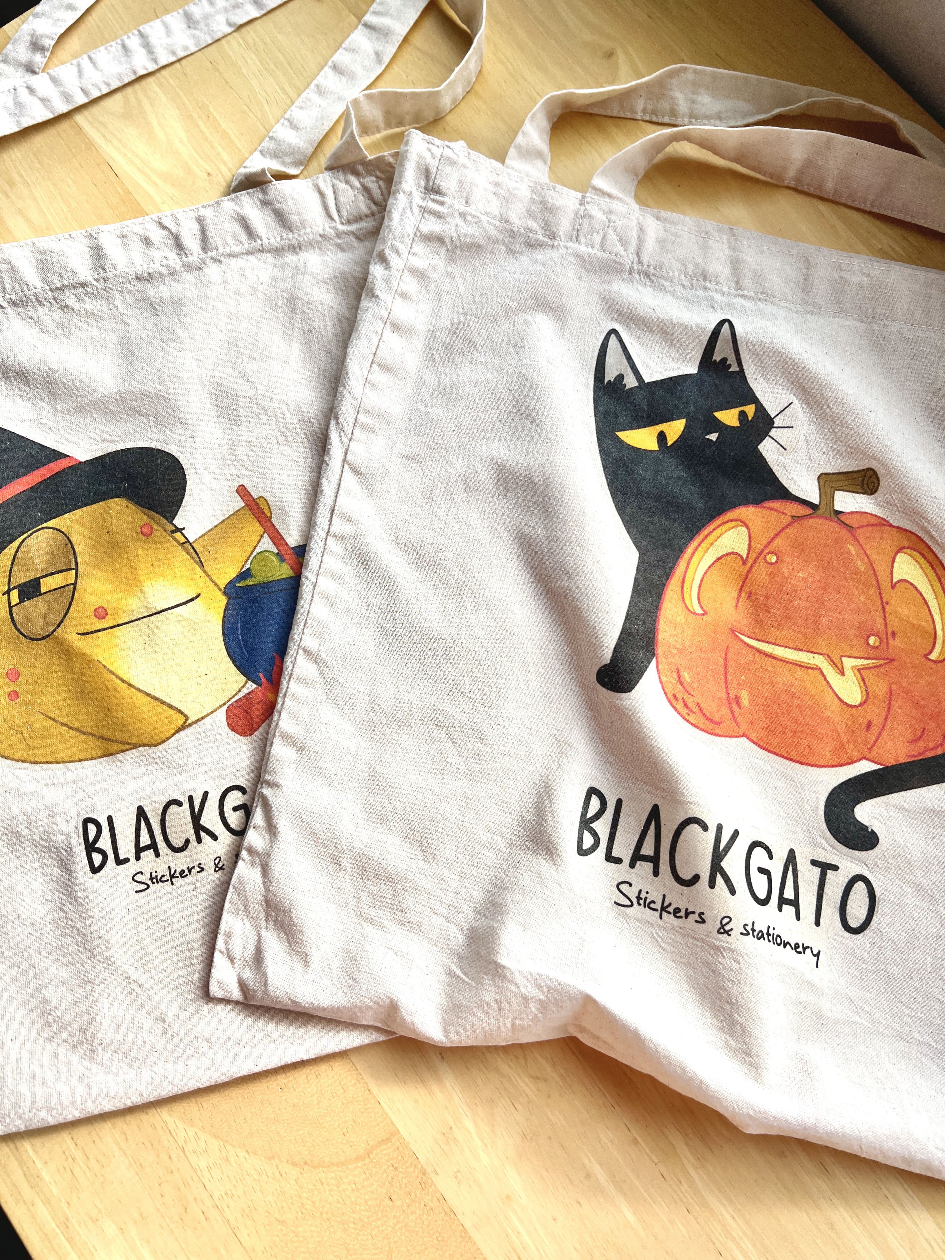 Two cotton tote bags featuring Halloween-themed designs from the Blackgato shop. One bag shows Toad, the yellow toad character, dressed as a witch stirring a cauldron. The other features Blackgato, the black cat, holding a carved pumpkin. Both totes lie flat on a wooden surface, surrounded by soft natural lighting.