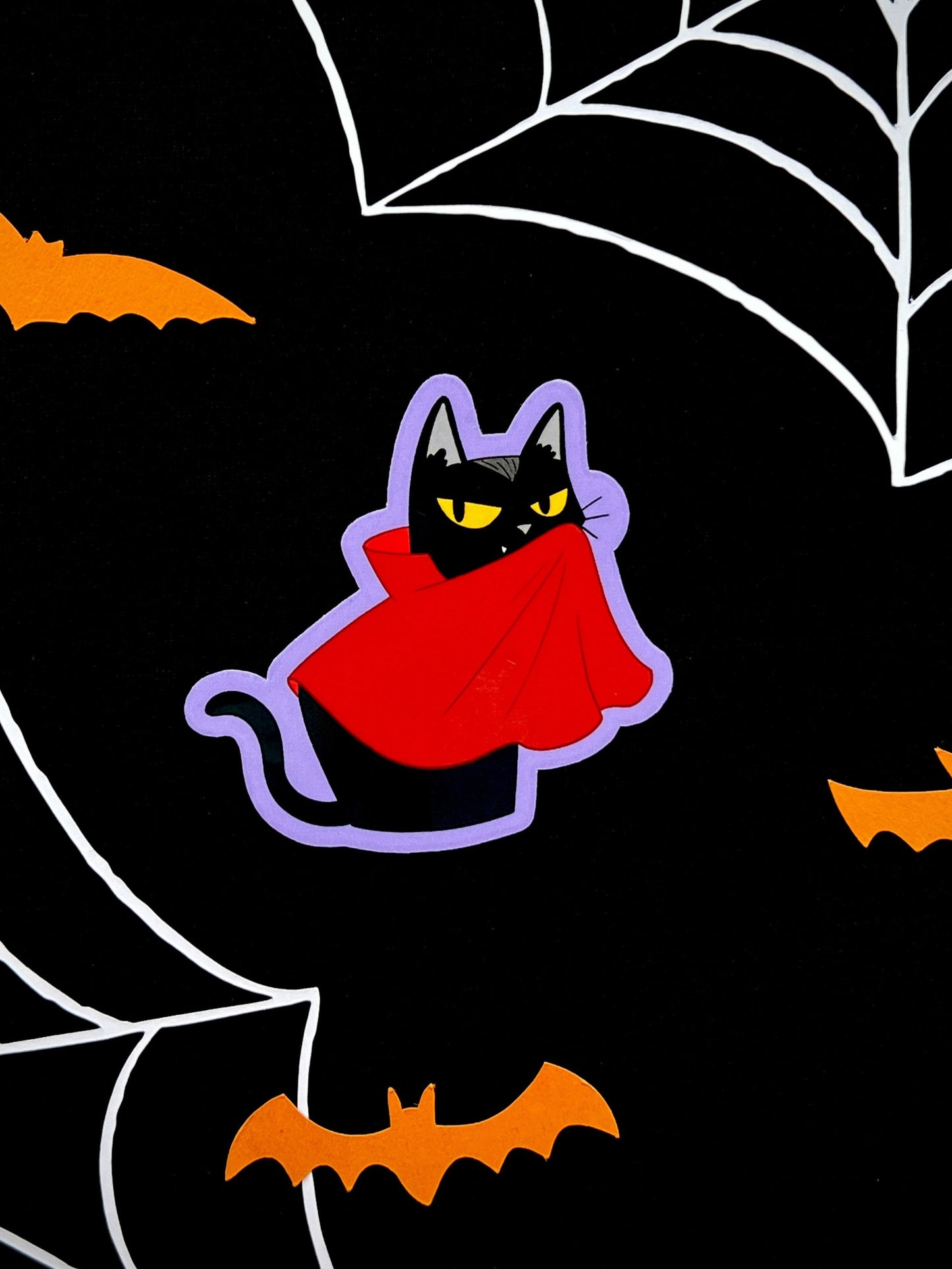 The image shows a die cut sticker of a black cat dressed as a vampire on a black background.