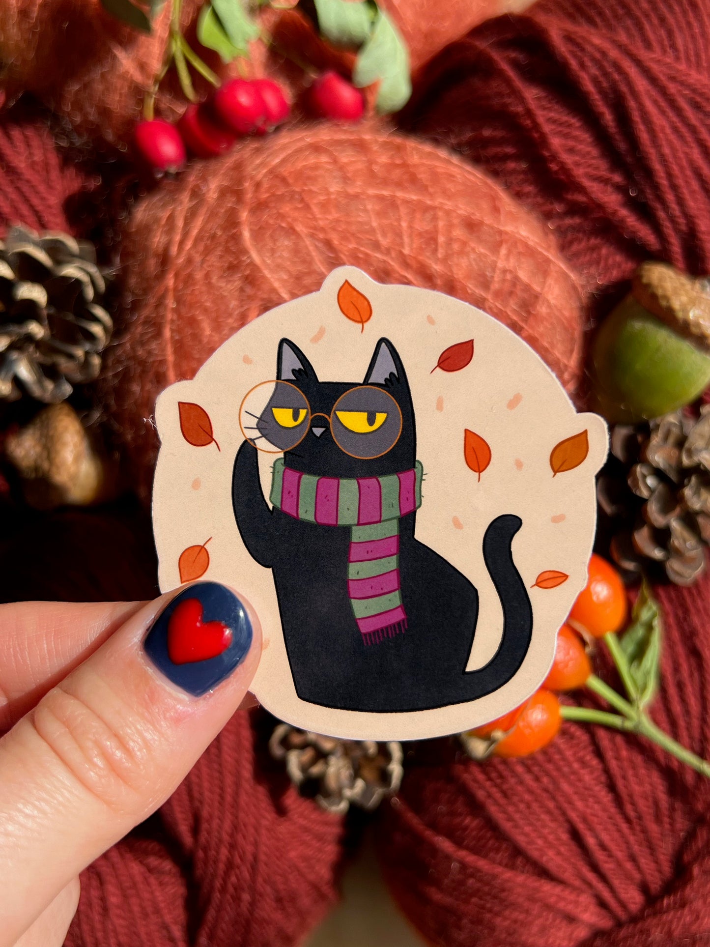 Blackgato autumn sticker featuring black cat, with hand for scale, on backdrop of cosy red yarn and pinecones.