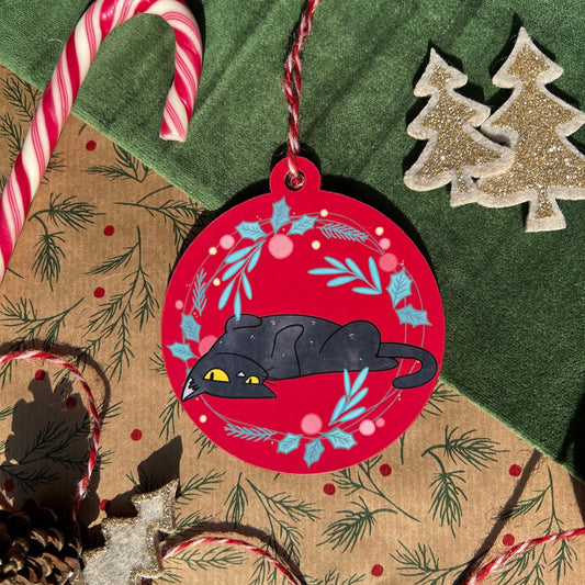 A festive flat-lay featuring a handmade Christmas bauble inspired by a traditional ornament shape. The bauble showcases Blackgato, a black cat, lying playfully on a vibrant red background adorned with a holly wreath. Surrounding it are holiday-themed props: candy canes, pinecones, and glittery tree decorations.