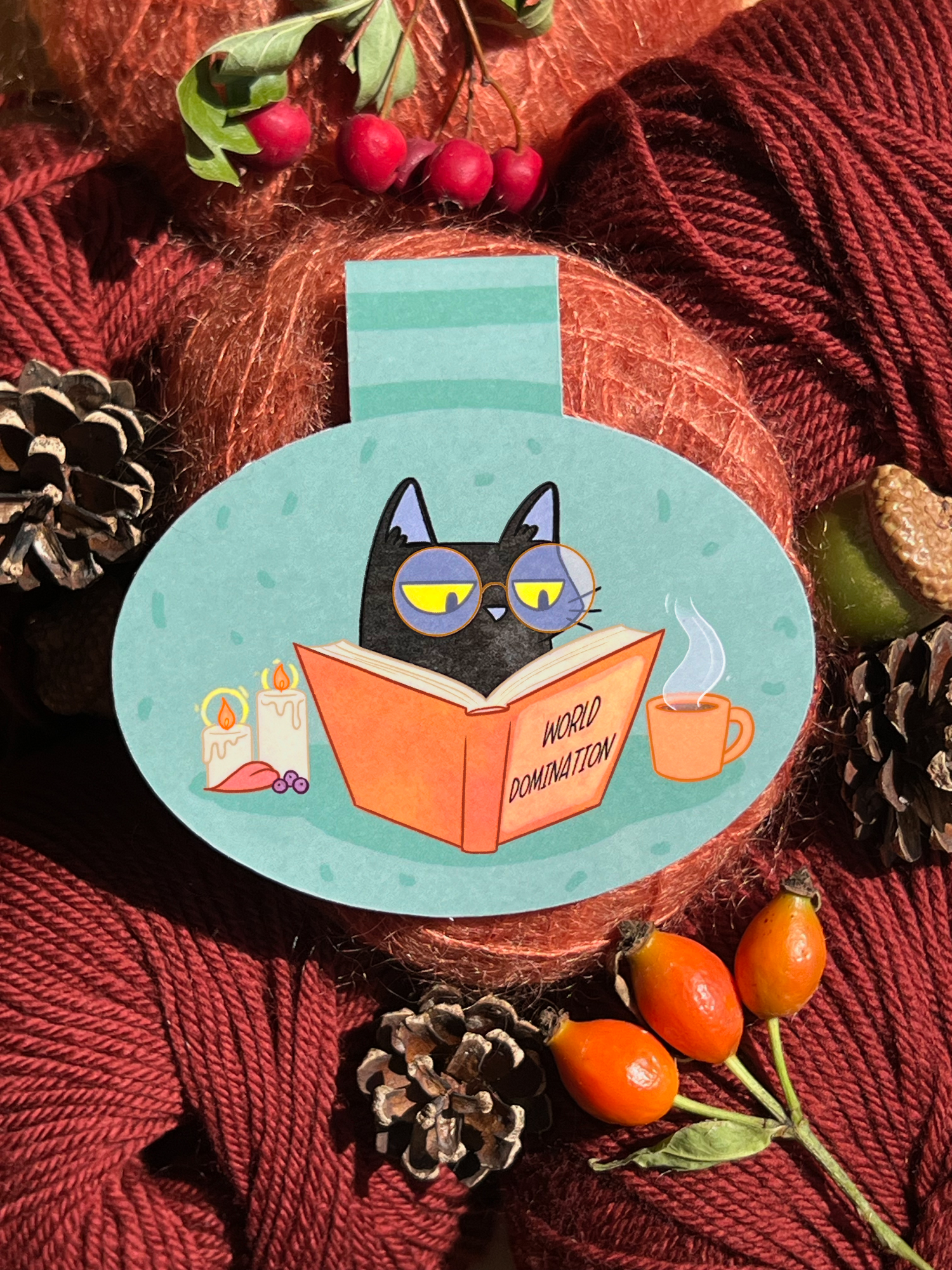 Blackgato magnetic bookmark with cosy red autumn themed backdrop. Illustration on the product shows black cat with glasses and coffee reading a book.