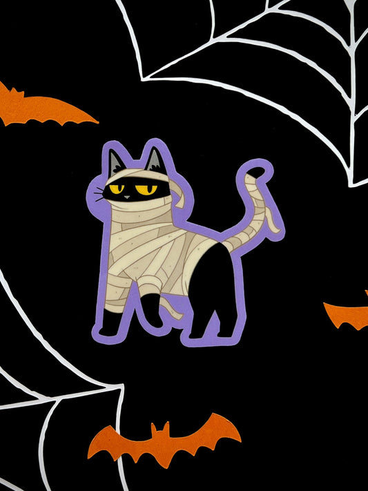 Die cut sticker of a black cat dressed as a mummy on a black background