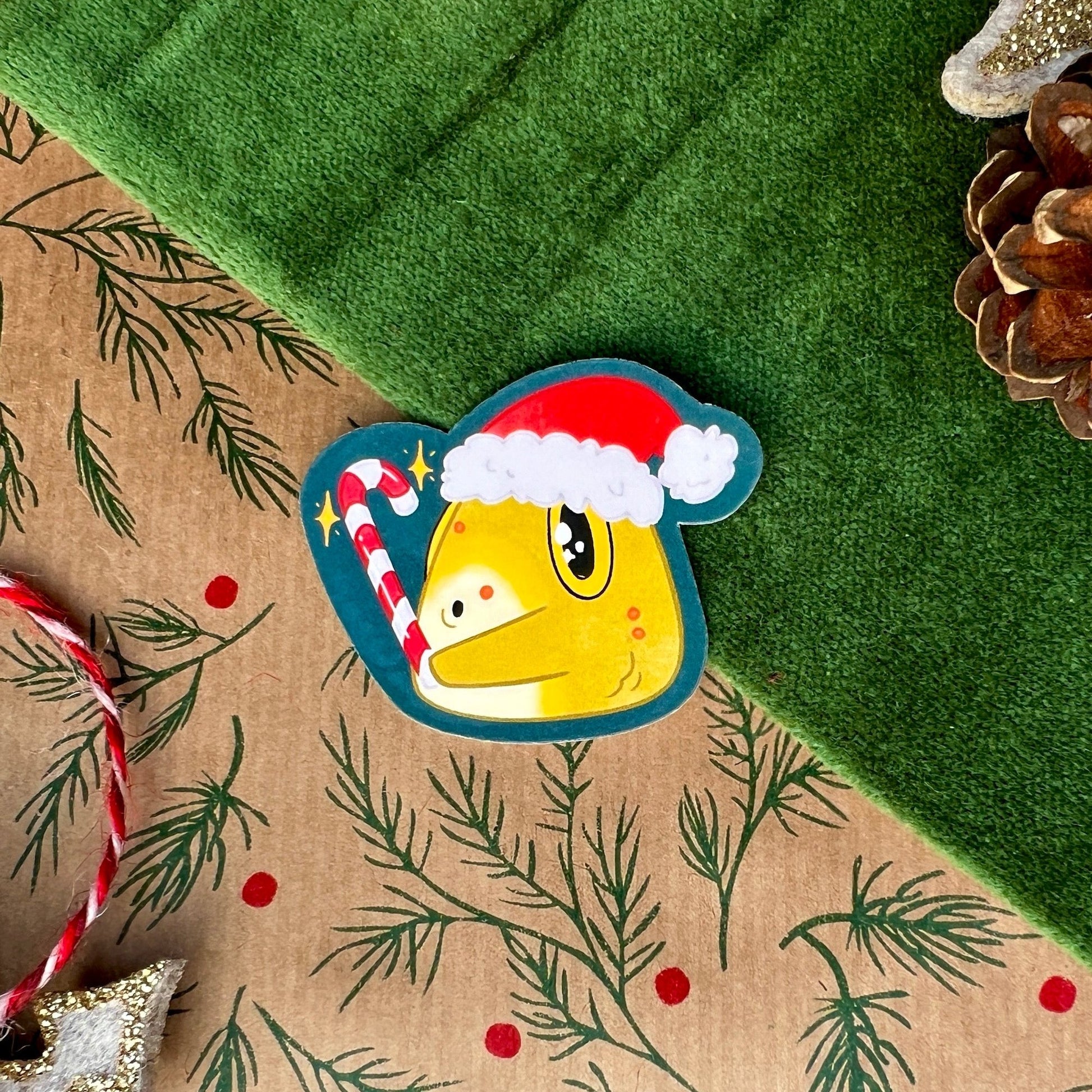 A sticker featuring a toad wearing a red Christmas hat, holding a candy cane that glows with magical sparkles. The sticker is displayed on a green festive background with pinecones and a candy cane nearby.