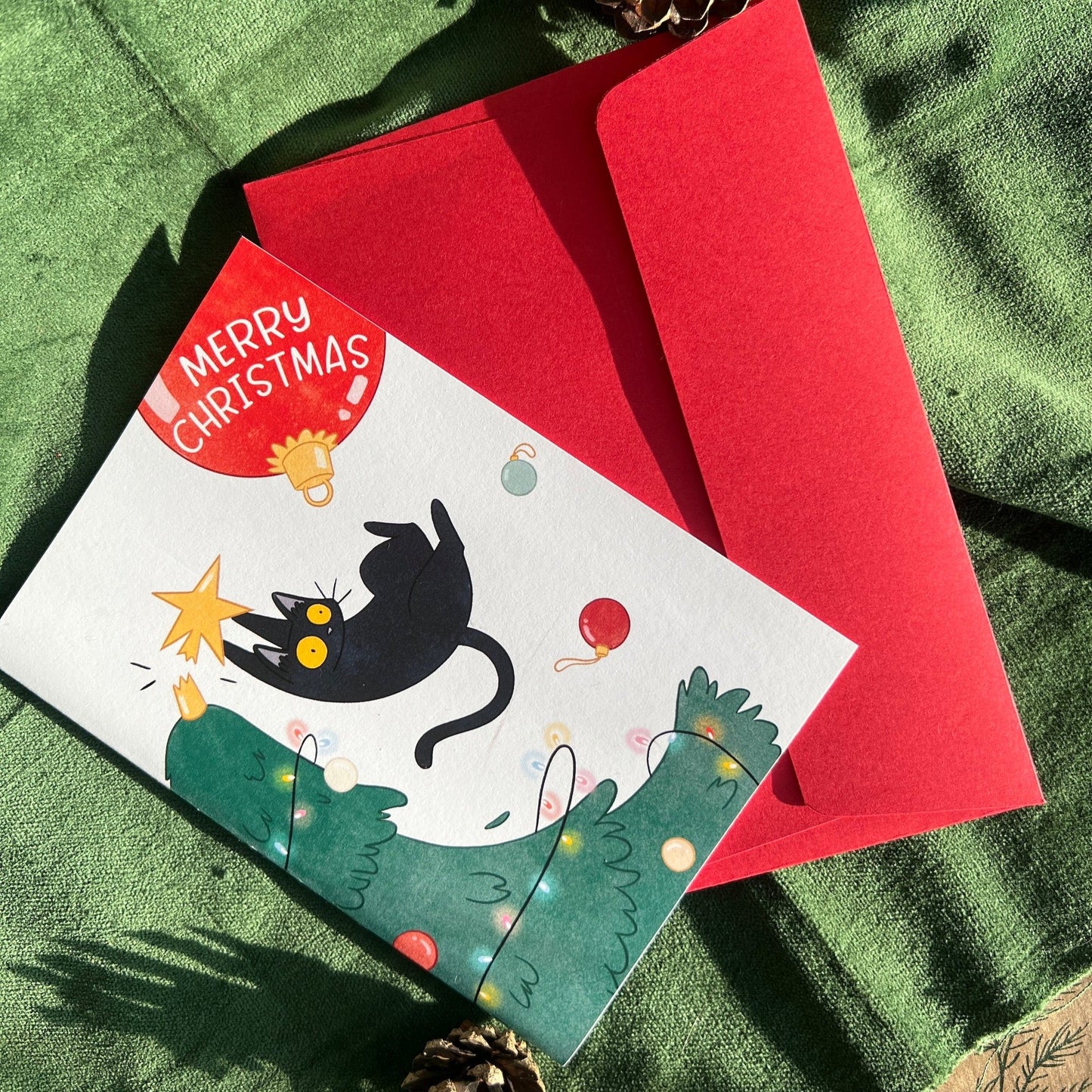 A Christmas greeting card featuring a playful illustration of a black cat leaping onto a Christmas tree, knocking the star topper off. The background includes a red ornament with the words "Merry Christmas," and the card is paired with a vibrant red envelope. The card is displayed on a green velvet surface with a pinecone as decoration.