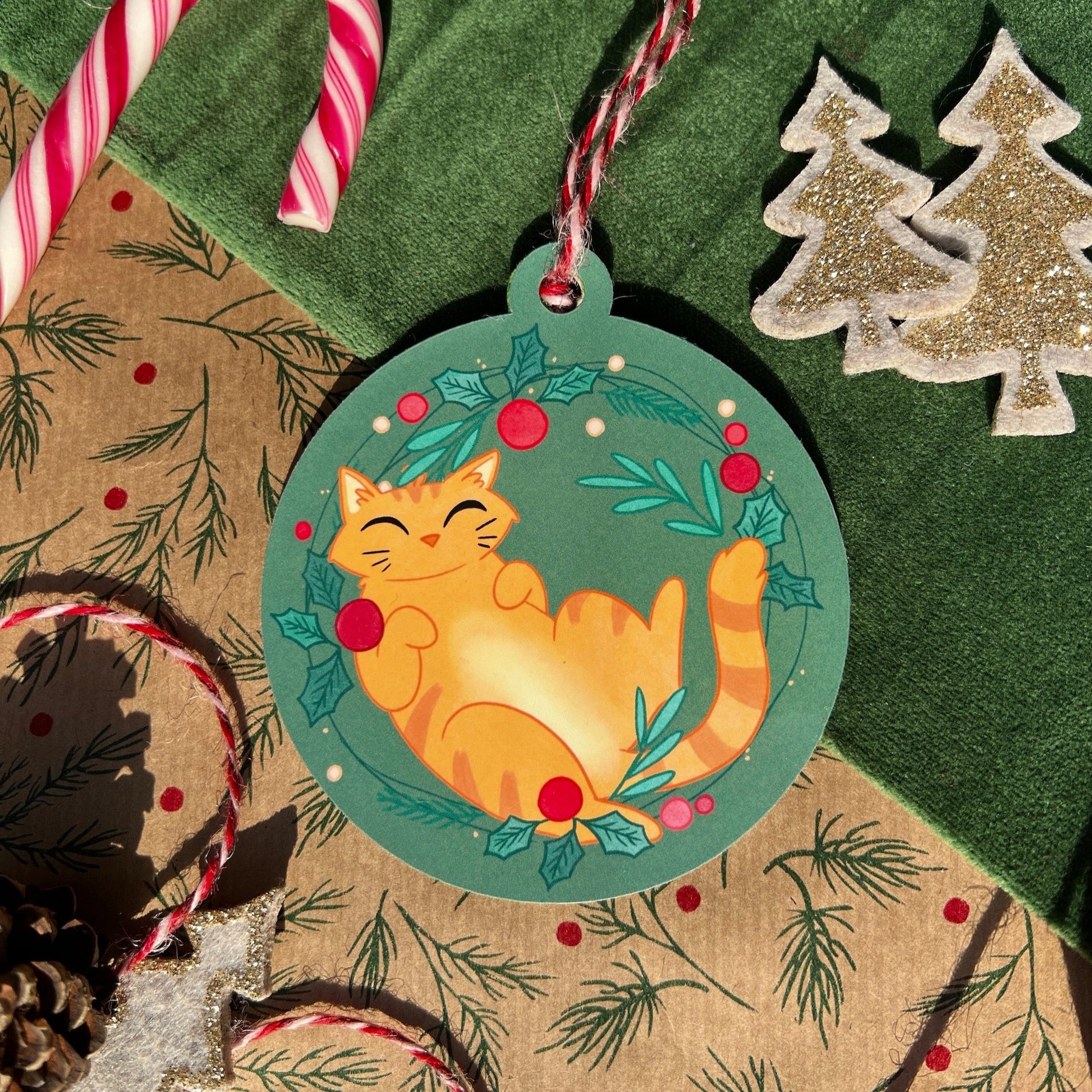 An image of a festive handmade Christmas bauble featuring Ginger, a cheerful orange cat, joyfully reclining on a green backdrop adorned with holly leaves and red berries. The bauble is placed on a decorative holiday setting with candy canes, pinecones, and glittery tree ornaments, exuding a cozy, seasonal charm.