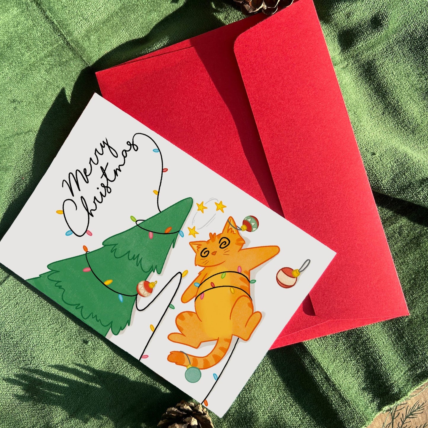 A cheerful Christmas card depicting an orange cat, Ginger, tangled in Christmas lights next to a tree, accompanied by a red envelope on a green festive background with pinecones.