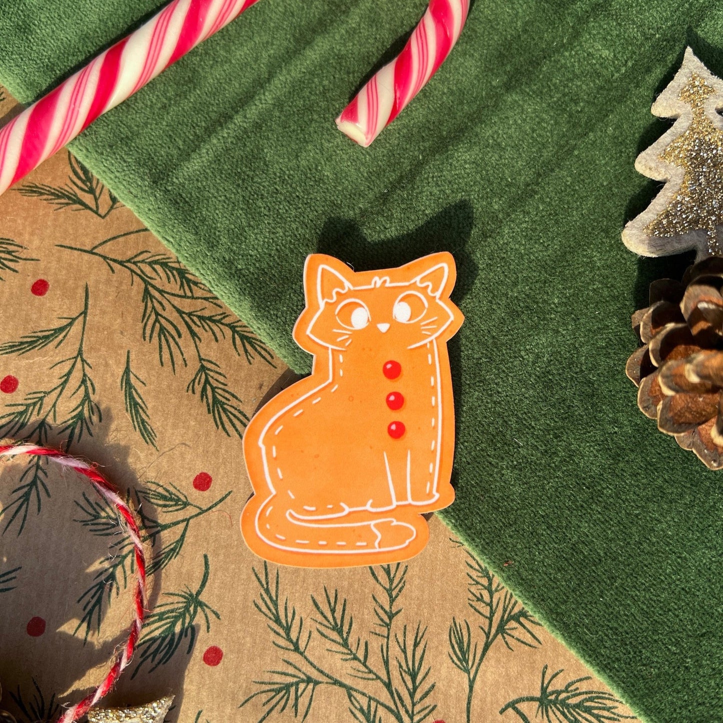 A gingerbread-style sticker shaped like a cat, featuring icing-like white outlines and red candy-like buttons, displayed on a green fabric background surrounded by festive decorations, including a candy cane and pinecones.