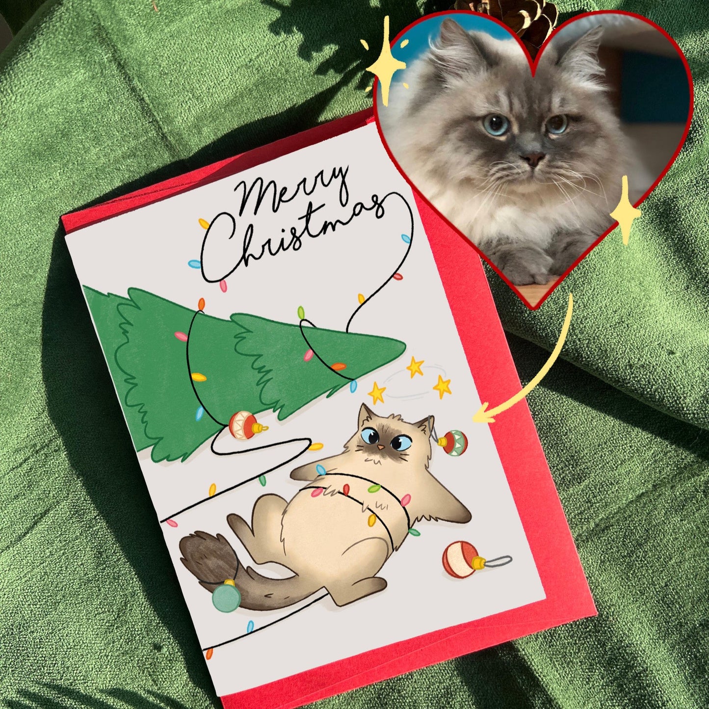 A personalized Christmas card featuring a hand-drawn illustration of a cat tangled in Christmas lights, styled after a customer's pet. The card lies on a green velvet surface, paired with a red envelope, surrounded by pinecones and festive decorations.