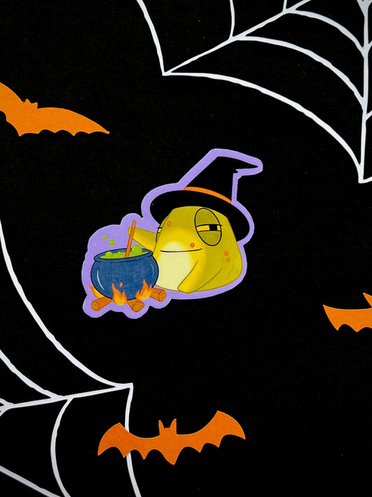 Sticker of a toad dressed as a witch. the toad is stirring a cauldron with a bubbly green liquid inside. The sticker is on a black background with Halloween decorations