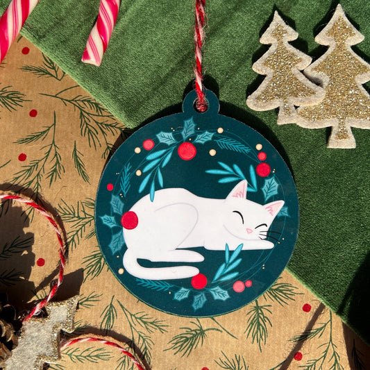 A handmade Christmas bauble featuring Snow, a white cat, peacefully curled up in the center of a festive green and red wreath design, with holly leaves and berries. The bauble lies on a decorative background with pine patterns, a candy cane, pinecone, and glittery mini Christmas tree ornaments.