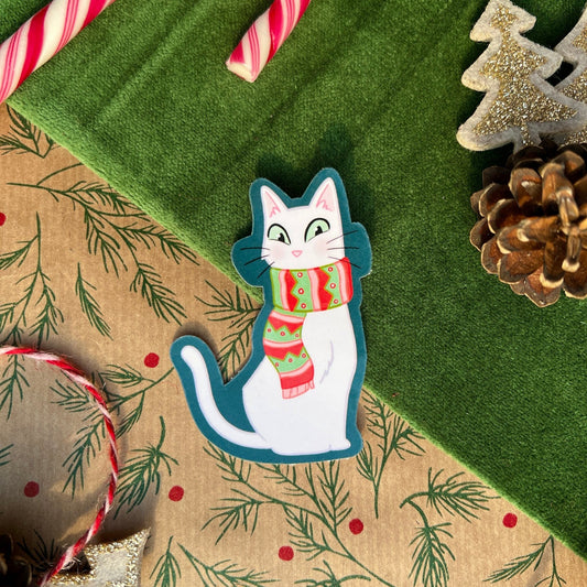 A sticker of a white cat named Snow, wearing a cozy, colourful scarf with festive red and green accents, placed on a green fabric background surrounded by holiday decorations, including a candy cane and pinecones.