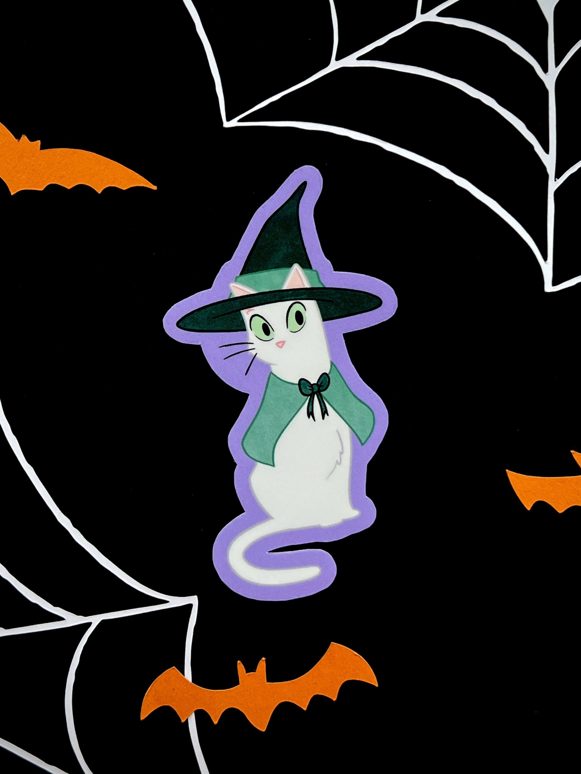 Die cut sticker of a white cat dressed as witch with a green cape, on a black background.