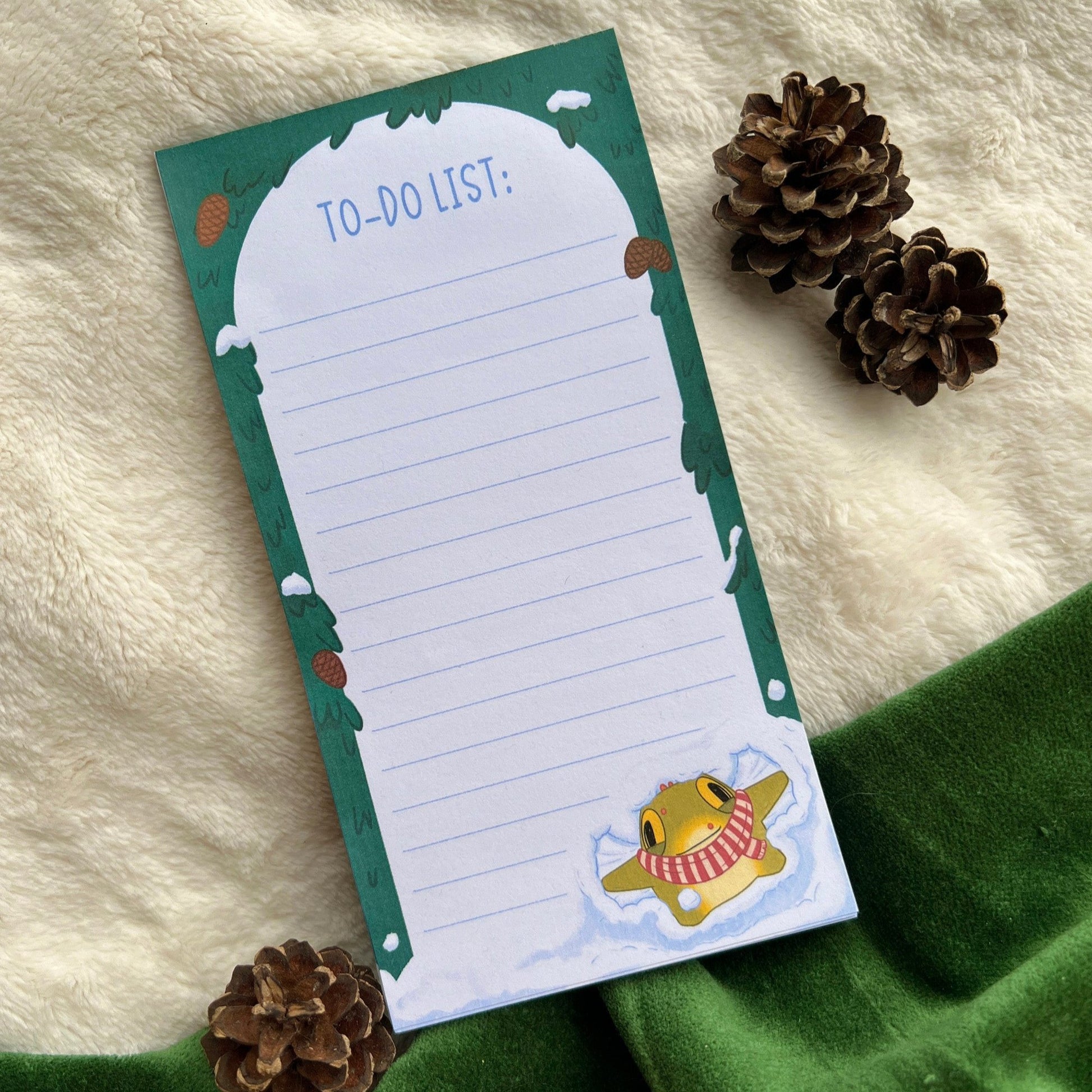 Winter-themed to-do list notepad with a snowy green forest border and a cute yellow character (Toad) at the bottom wearing a red scarf, surrounded by snow, placed on a soft beige blanket with pinecones nearby.