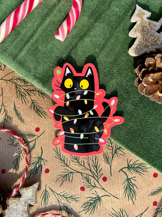 A festive sticker featuring a black cat tangled in colorful Christmas lights, with a red border accentuating the playful design. The sticker is set on a holiday-themed background with candy canes, pinecones, and a glittering tree ornament for a cozy seasonal vibe.