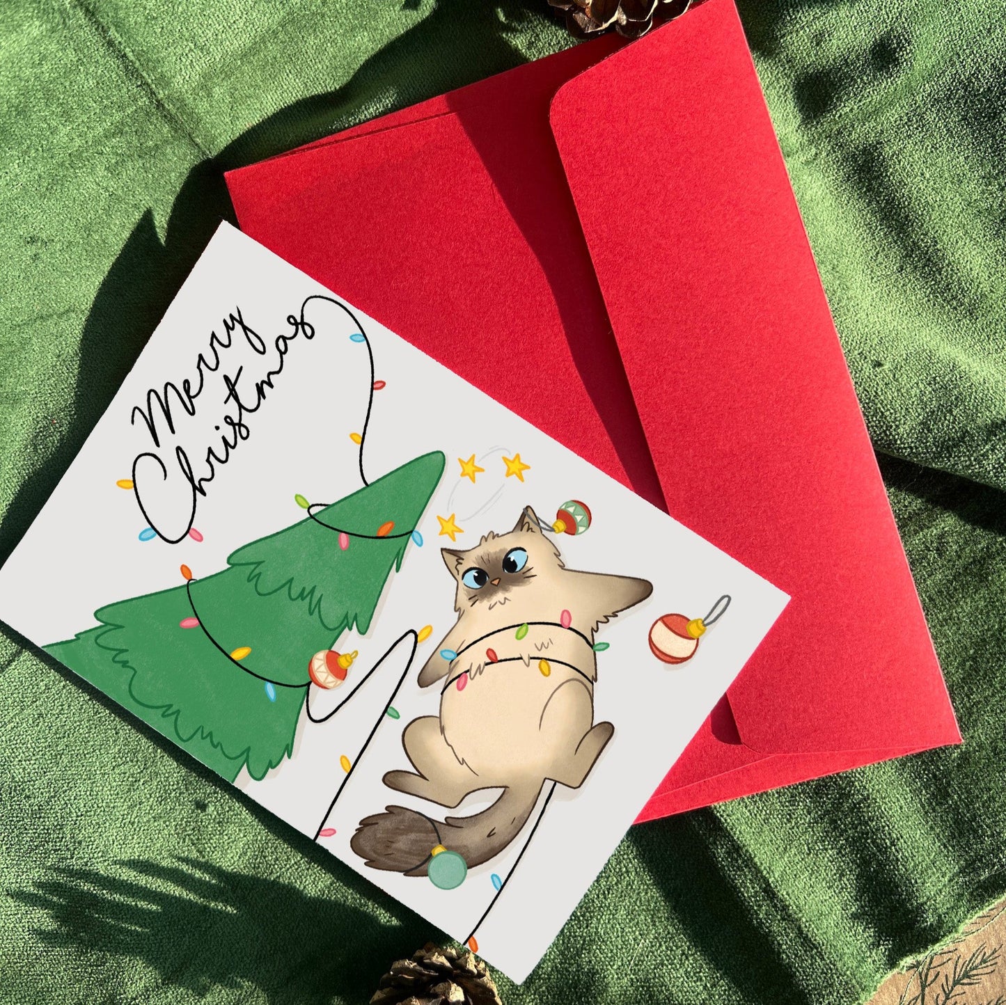 A Christmas greeting card featuring a playful illustration of a fluffy Ragdoll cat tangled in colorful string lights. The mischievous cat leaps onto a Christmas tree, toppling ornaments and the star topper. The card is paired with a vibrant red envelope, displayed on green velvet with a pinecone.