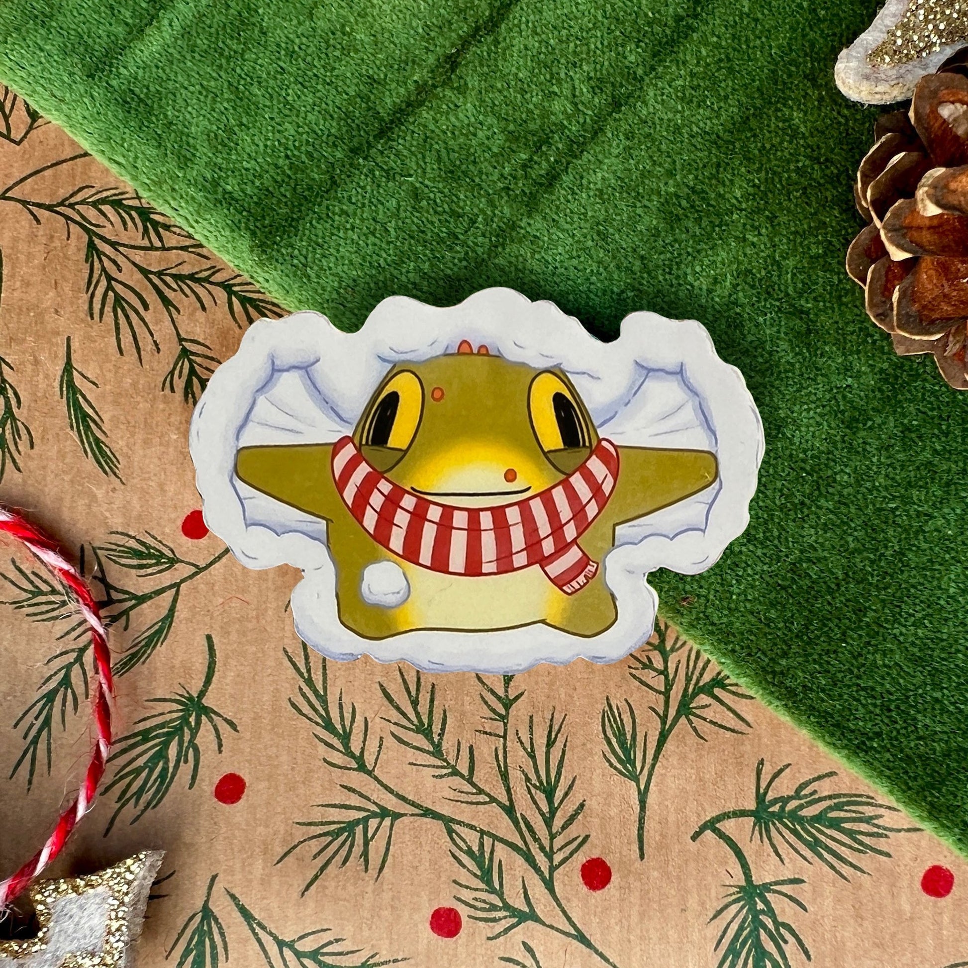 A festive sticker featuring Toad, a cheerful yellow toad, wearing a red-and-white striped scarf and creating a snow angel in fluffy white snow. The sticker is displayed on a green velvet background with a candy cane, pinecone, and Christmas-themed decorations.